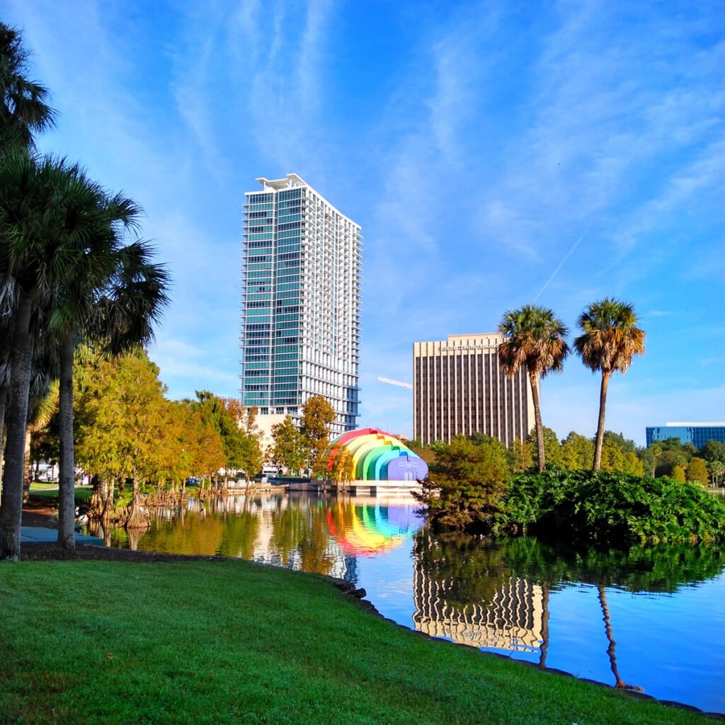 Ein Stadtpark mit Palmen und einem Teich, der nahe gelegene Gebäude und eine Regenbogen-farbige Struktur unter einem blauen Himmel reflektiert.