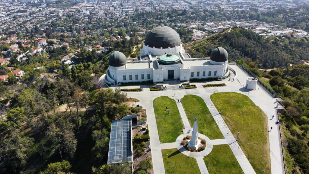 Luftaufnahme des Griffith Observatory mit seinen Kuppeln, umliegenden Wegen und grünen Rasenflächen, im Hintergrund mit Blick auf die Stadtlandschaft von Los Angeles.