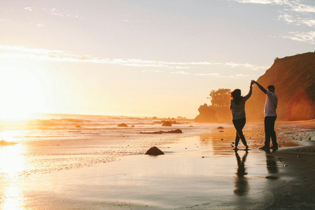 Zwei Menschen tanzen bei Sonnenuntergang an einem Sandstrand, im Hintergrund sind Wellen, Felsen und Klippen zu sehen.