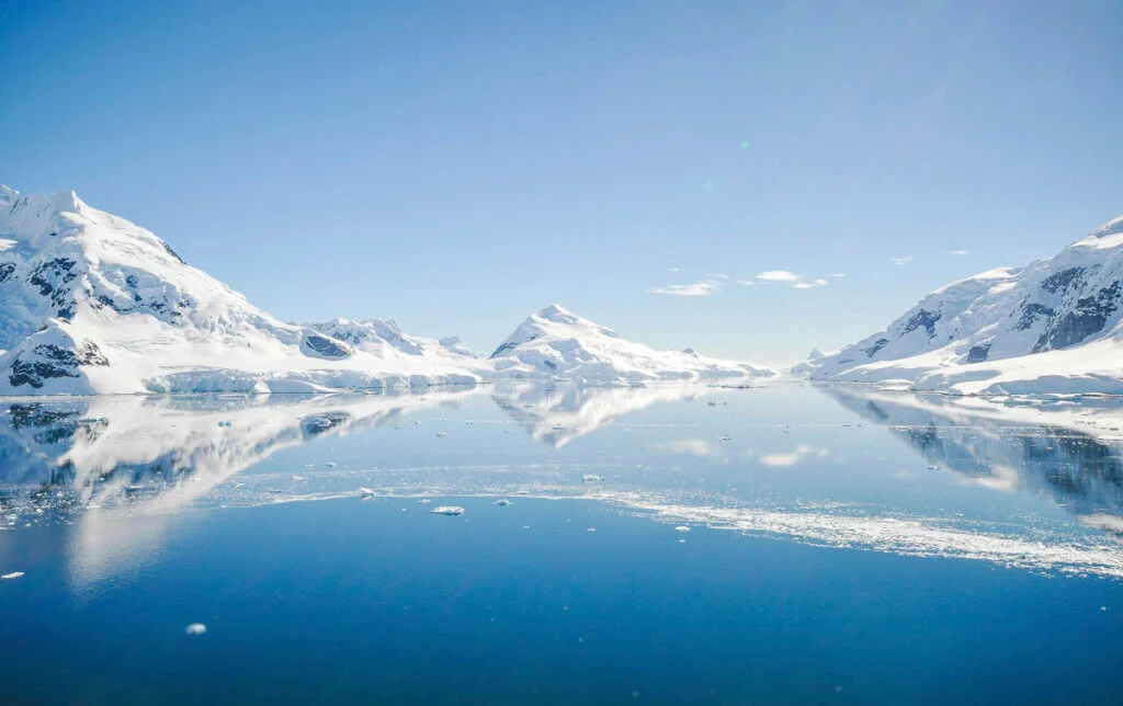 Schneebedeckte Berge spiegeln sich in einem ruhigen, eisigen Gewässer unter einem klaren blauen Himmel.