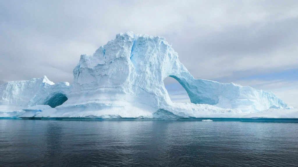 Großer Eisberg mit bogenförmigen Öffnungen, der in ruhigem, dunklem Wasser unter bewölktem Himmel schwimmt.