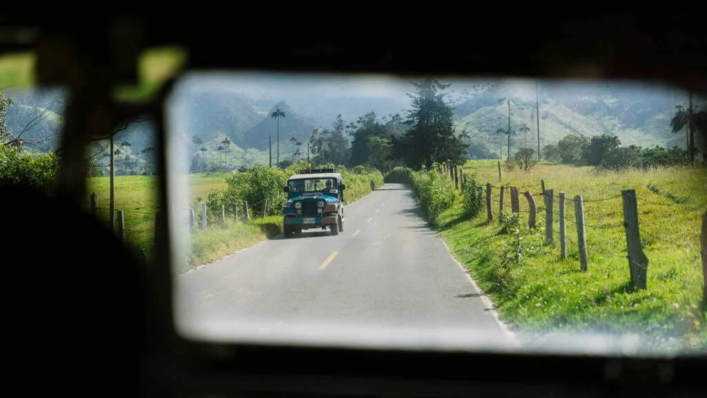 Ein blau-weißer Oldtimer-Jeep fährt eine von Gras und Zaunpfählen gesäumte Landstraße entlang, im Hintergrund sind Berge zu sehen.