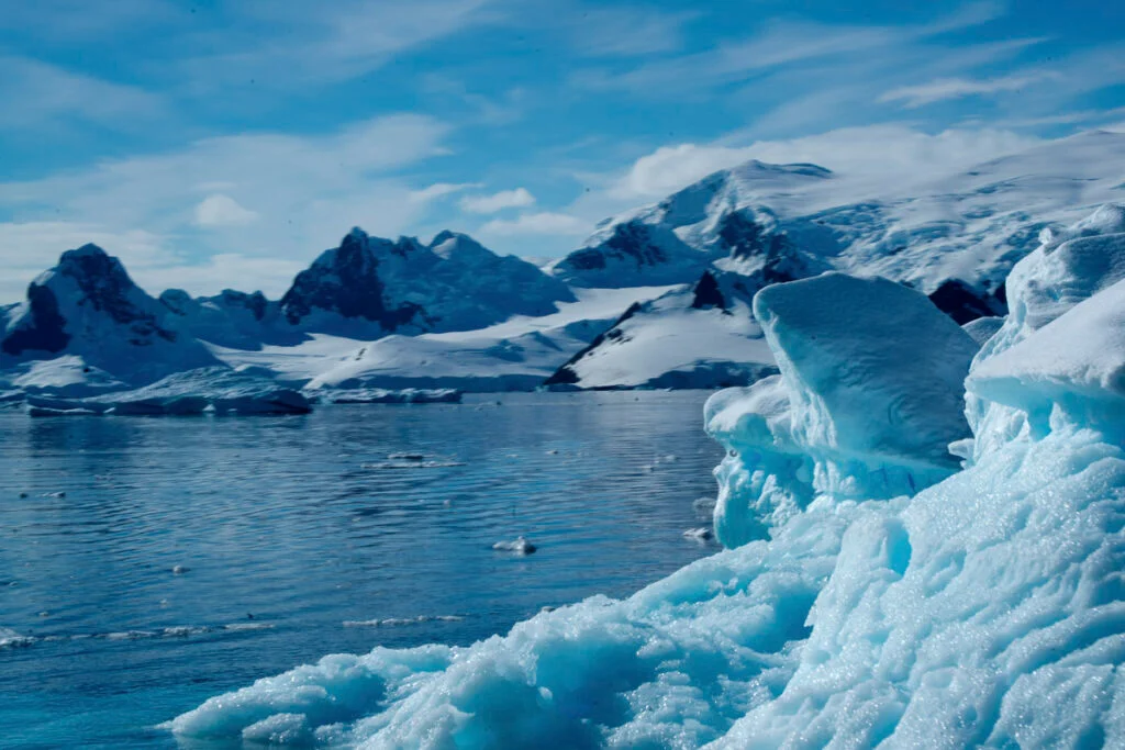 Schneebedeckte Berge und Gletscher erheben sich über einem ruhigen Gewässer unter einem blauen Himmel mit vereinzelten Wolken.