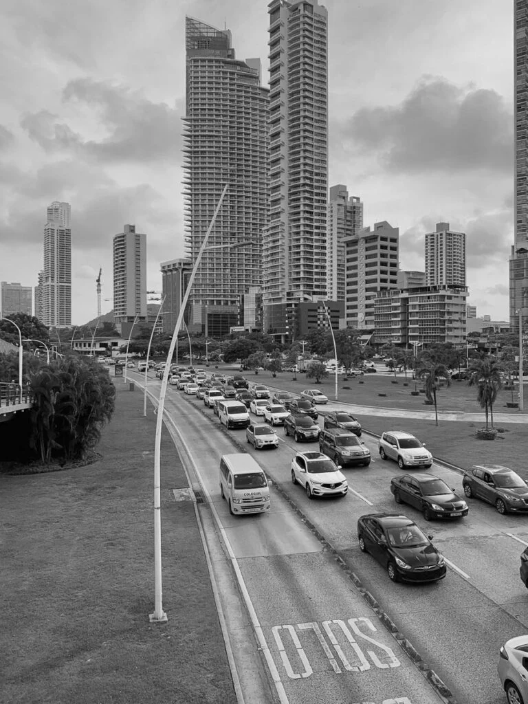 Schwarz-Weiß-Foto einer belebten Stadtstraße mit mehreren Fahrspuren im Verkehr, umgeben von hohen modernen Gebäuden und bewölktem Himmel.
