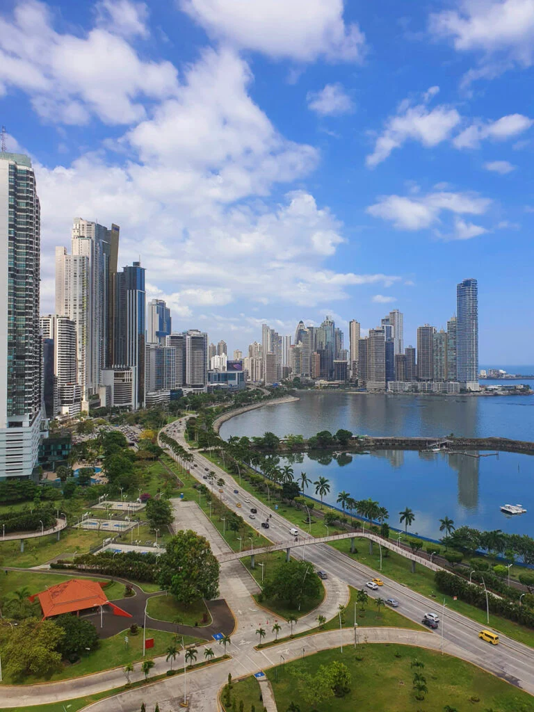 Eine Stadtlandschaft aus modernen Hochhäusern säumt eine geschwungene Uferpromenade unter einem teilweise bewölkten Himmel, mit einem Park und stark befahrenen Straßen im Vordergrund.