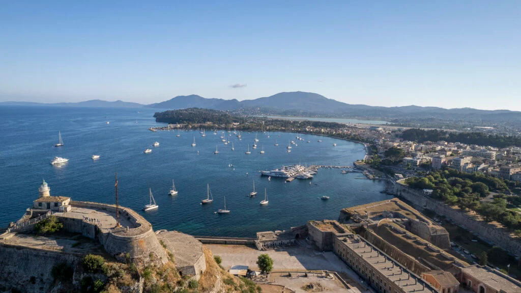 Luftaufnahme einer Küstenstadt mit einem Hafen voller Boote, historischen Befestigungsanlagen im Vordergrund und Bergen im Hintergrund unter klarem Himmel.