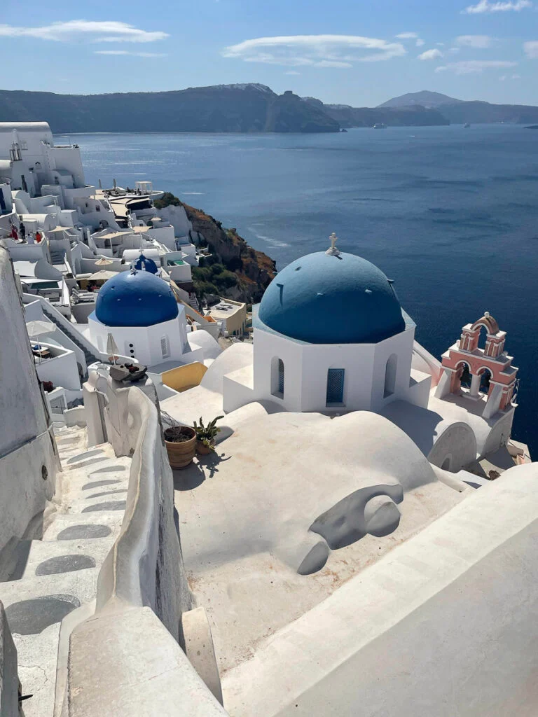 Weiße Gebäude mit blauen Kuppeln überblicken das Ägäische Meer in Santorini, Griechenland, mit Klippen und fernen Inseln, die unter einem teilweise bewölkten Himmel sichtbar sind.