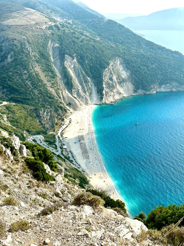 Luftaufnahme eines Sandstrandes mit türkisfarbenem Wasser, gesäumt von steilen Klippen und grünen Hügeln, mit Sonnenliegen und Sonnenschirmen entlang der Küste.