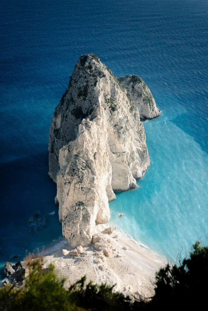 Eine große Felsformation erhebt sich aus türkisblauem Wasser neben einem Sandstrand, von oben betrachtet mit grünem Blattwerk im Vordergrund.
