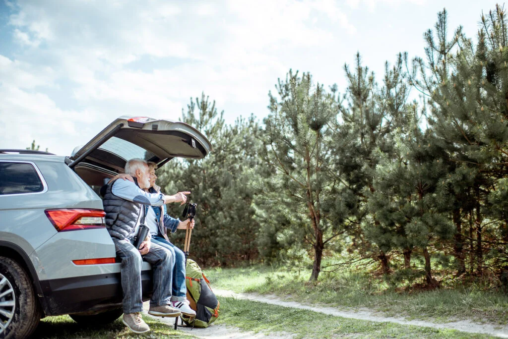Zwei ältere Erwachsene sitzen an einem sonnigen Tag am offenen Kofferraum eines SUV, halten Wanderstöcke in der Hand und blicken auf einen Kiefernwald.