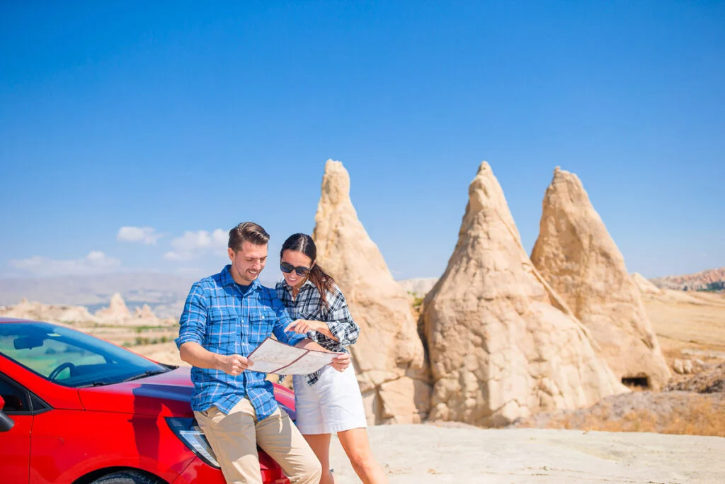 Ein Mann und eine Frau stehen neben einem roten Auto und betrachten eine Karte mit einzigartigen Felsformationen und einem klaren blauen Himmel im Hintergrund.