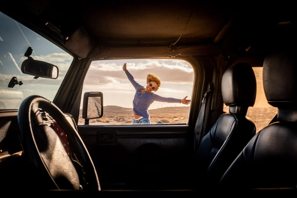 Ein Kind mit Sonnenbrille und blauem Hemd springt mit weit geöffneten Armen vor einem geparkten Auto in einer Wüstenlandschaft, gesehen durch das offene Fenster des Autos.
