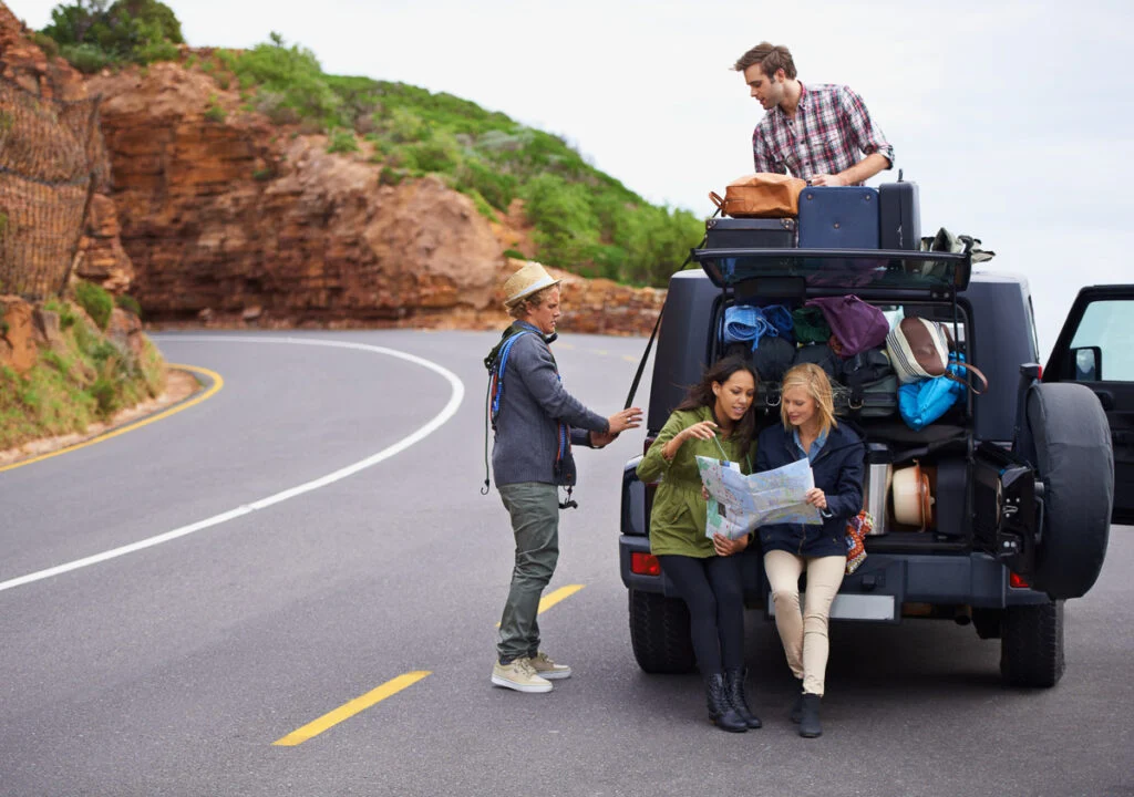 Vier Personen sind auf einem Roadtrip und werden mit ihrem SUV am Straßenrand angehalten. Zwei Frauen schauen auf eine Karte, während ein Mann sein Gepäck auf dem Dach stapelt und ein anderer in der Nähe steht.