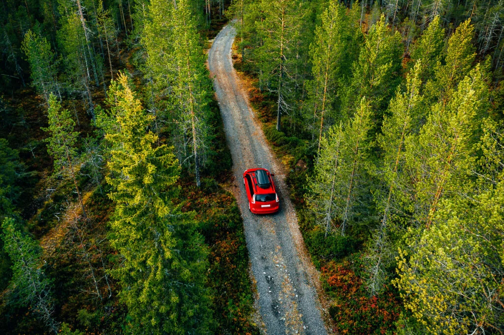 Ein rotes Auto fährt von oben betrachtet eine schmale Schotterstraße entlang, umgeben von dichtem grünen Wald.