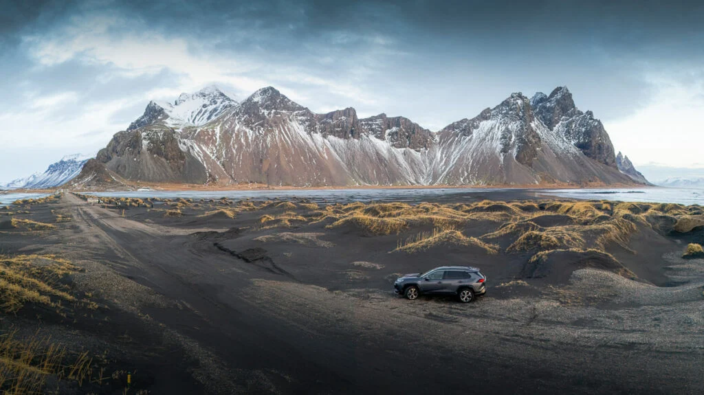 Ein schwarzer SUV parkt auf einer sandigen, schwarzen Landschaft mit schneebedeckten Bergen im Hintergrund unter bewölktem Himmel.