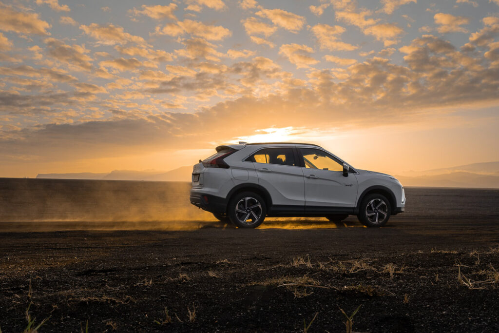 Ein weißer SUV fährt bei Sonnenuntergang auf einer unbefestigten Straße, Staub steigt um seine Räder auf und am Himmel sind Wolken zu sehen.