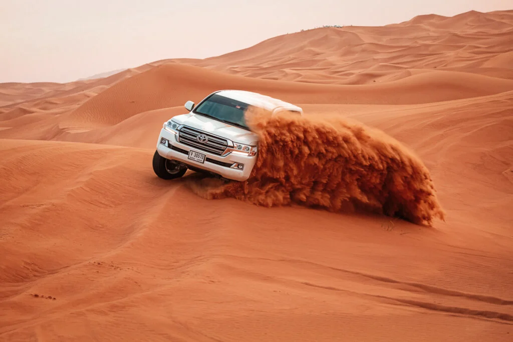 Ein weißer SUV fährt schnell durch orangefarbene Sanddünen und wirbelt dabei eine große Sandwolke hinter seinen Reifen auf.