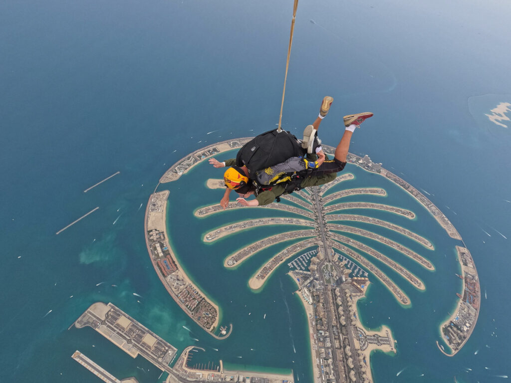 Eine Person beim Fallschirmspringen über der Palm Jumeirah, einer künstlichen Insel in Form einer Palme, umgeben von blauem Wasser in Dubai.
