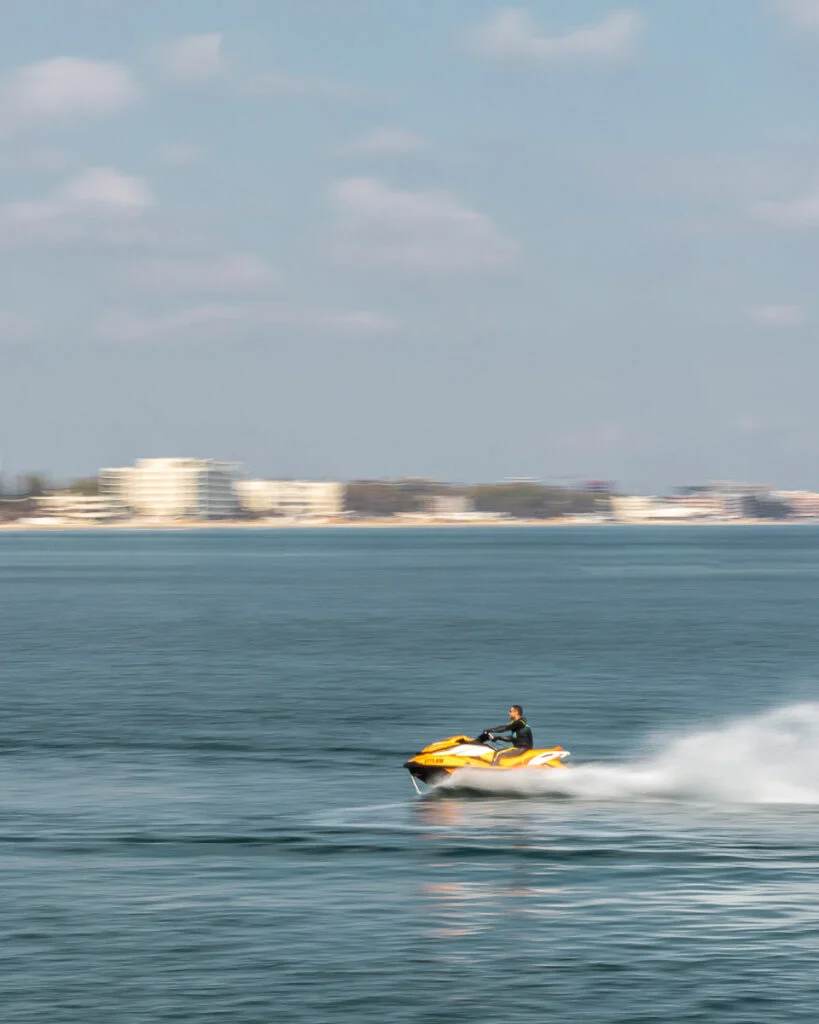 Person, die mit einem gelben Jetski über ruhiges blaues Wasser fährt, mit einer verschwommenen Stadtküste und Gebäuden im Hintergrund unter einem teilweise bewölkten Himmel.