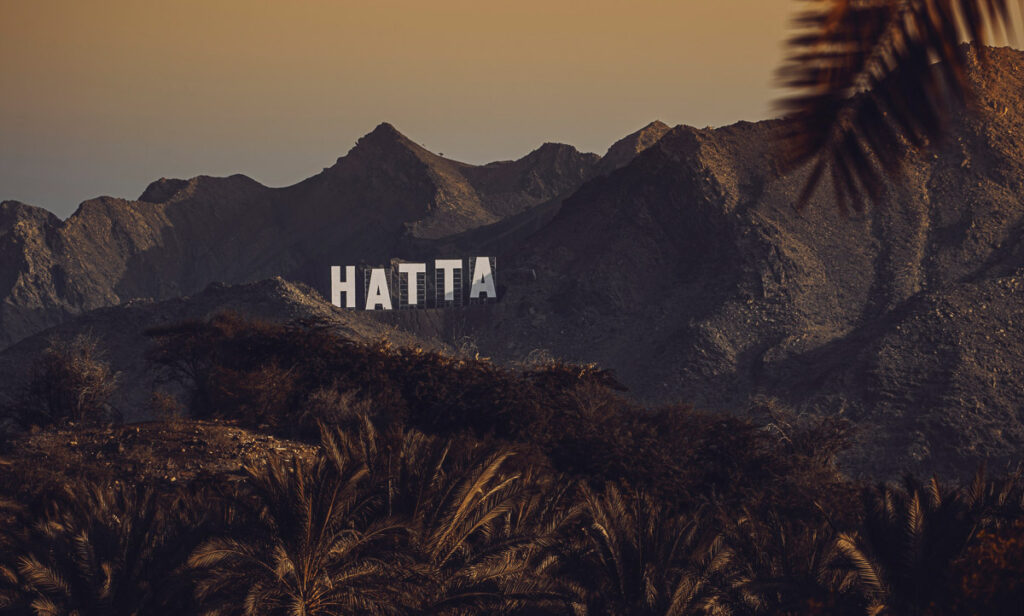 Auf einem felsigen Hügel steht ein großes weißes Schild mit der Aufschrift „HATTA“, mit Palmen im Vordergrund und schroffen Bergen im Hintergrund bei Sonnenuntergang.