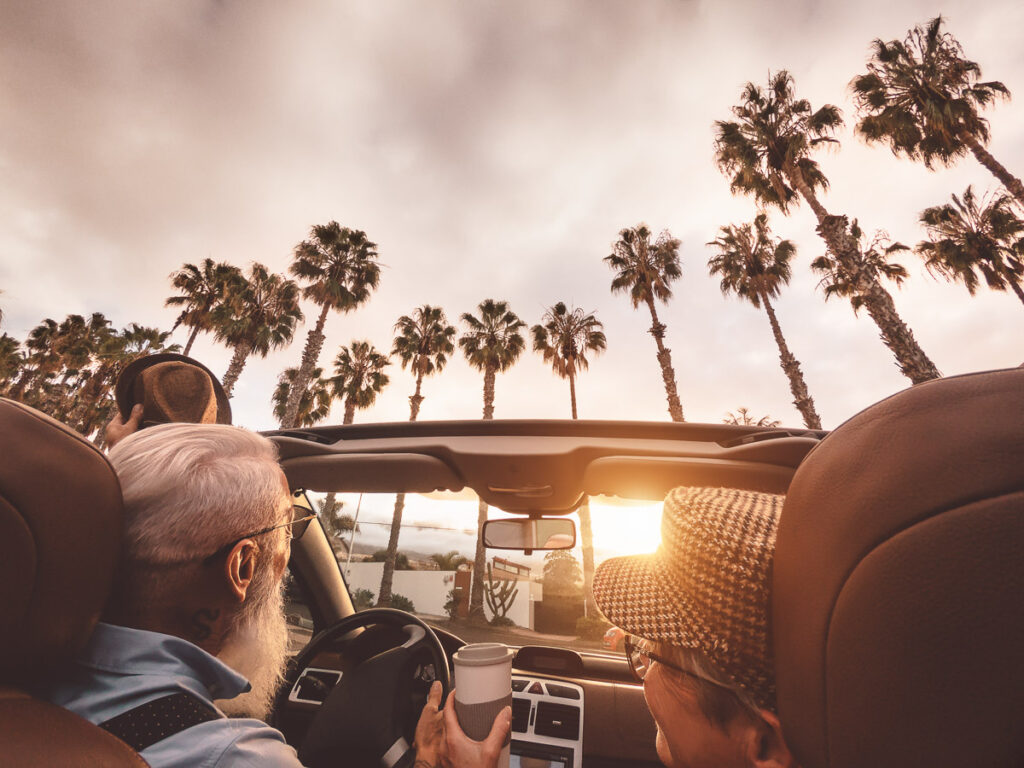Zwei Personen fahren in einem Cabrio mit offenem Verdeck vorbei an Palmen bei Sonnenuntergang. Einer hält eine Tasse und beide tragen Hüte. Der Himmel ist teilweise bewölkt.