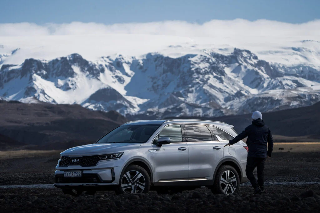 Eine Person mit Jacke steht neben einem silbernen SUV, der auf felsigem Gelände geparkt ist, im Hintergrund schneebedeckte Berge.