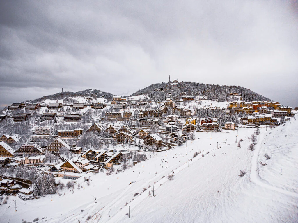 Schneebedeckte Gebäude und Bäume an einem Hang unter bewölktem Himmel, im Vordergrund Menschen, die Ski fahren oder auf den Pisten spazieren gehen.