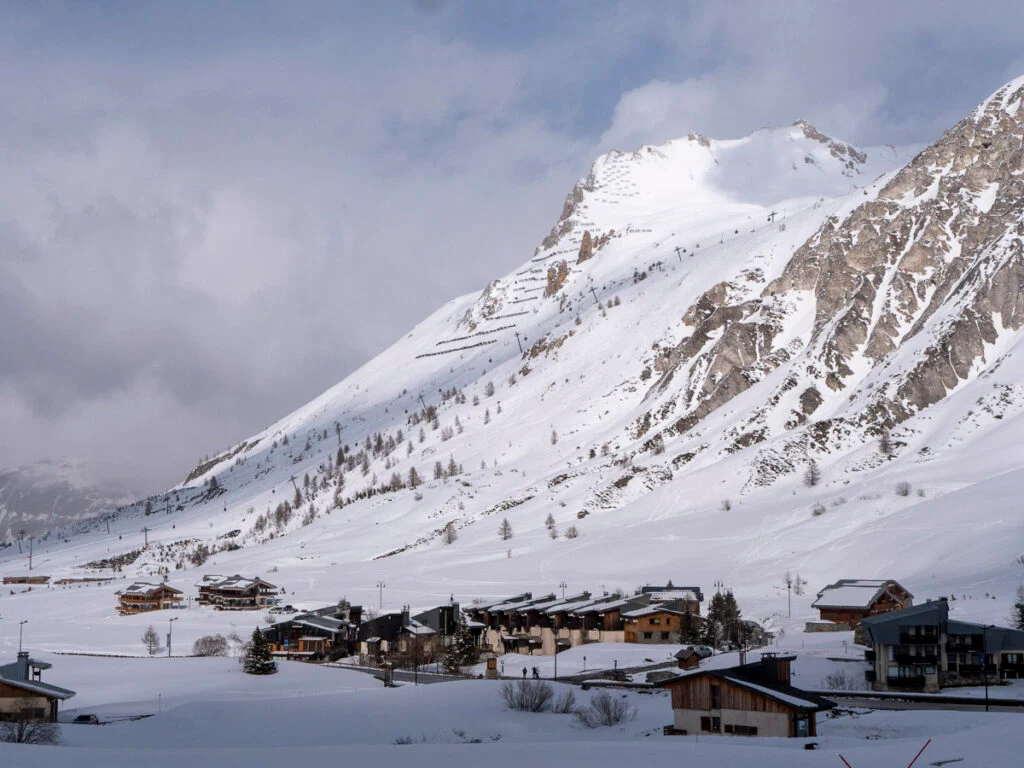 Ein schneebedeckter Berg mit vereinzelten Bäumen erhebt sich hinter einem kleinen Alpendorf aus Holzgebäuden unter bewölktem Himmel.