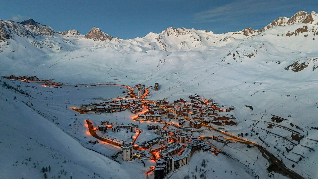 Luftaufnahme eines schneebedeckten Bergferienortes in der Abenddämmerung, mit beleuchteten Straßen, die sich zwischen Gebäuden schlängeln, und im Hintergrund sind die umliegenden Gipfel zu sehen.