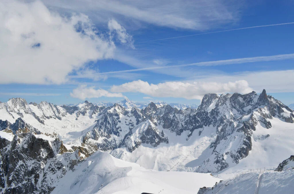 Schneebedeckte Berggipfel unter blauem Himmel mit vereinzelten Wolken und sichtbaren Kondensstreifen.