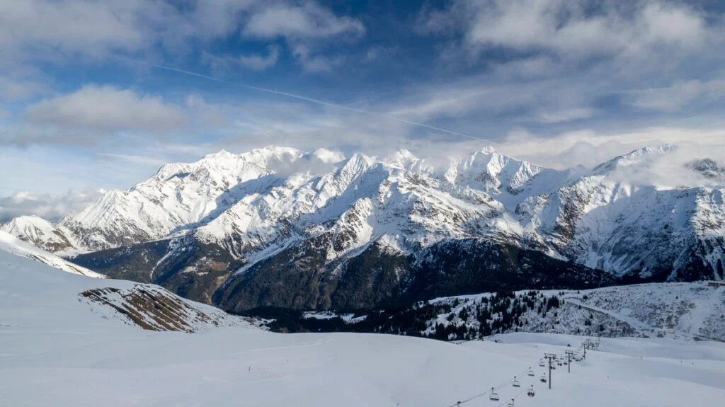 Schneebedeckte Berge unter einem teilweise bewölkten Himmel mit einem Skilift in der unteren rechten Ecke.