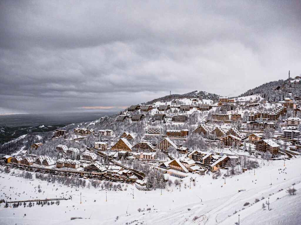 Verschneites Bergdorf mit mehreren Gebäuden im Chalet-Stil unter bewölktem Himmel, umgeben von schneebedeckten Hängen und Bäumen.