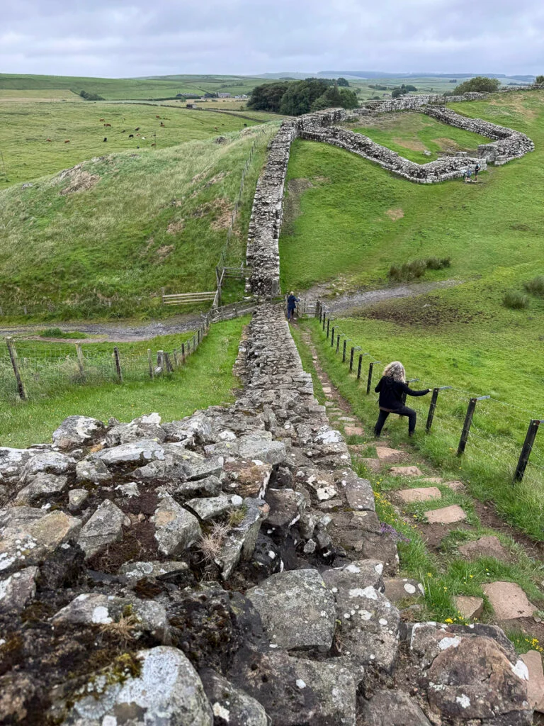Eine Person erklimmt Steinstufen entlang des Hadrianswalls, der sich unter bewölktem Himmel über eine Graslandschaft erstreckt.