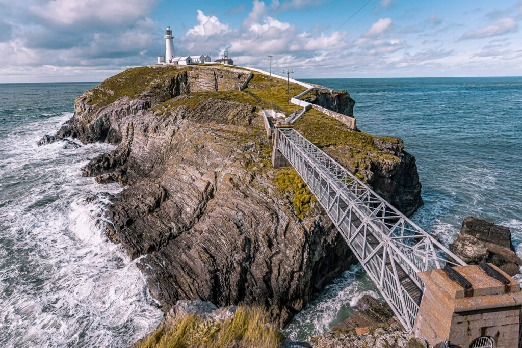 Eine metallene Fußgängerbrücke führt zu einer felsigen Insel mit einem Leuchtturm und Gebäuden, umgeben von Meereswellen unter einem teilweise bewölkten Himmel.