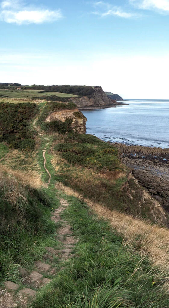 Ein schmaler Steinpfad schlängelt sich entlang grasbewachsener Küstenklippen und bietet Blick auf das ruhige blaue Meer und die felsige Küste unter einem teilweise bewölkten Himmel.