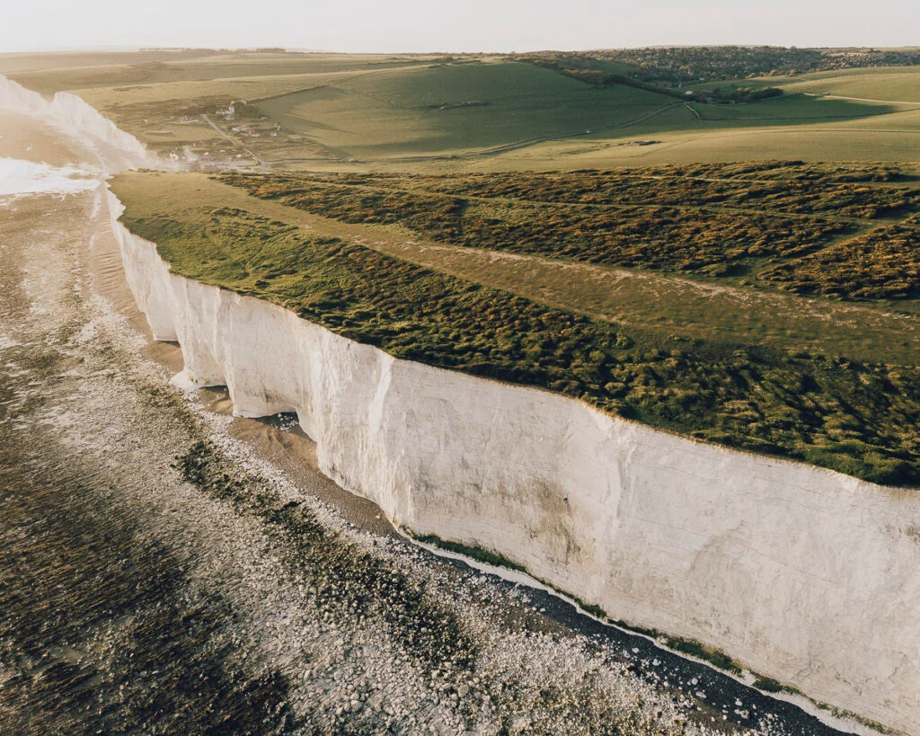 Luftaufnahme von hohen weißen Kreidefelsen, die auf das Meer treffen, mit grünen Grasfeldern oben und einer felsigen Küste unten.