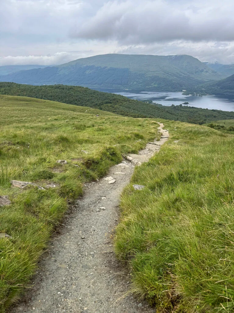 Schmaler Schotterweg durch grasbewachsene Hügel, der unter bewölktem Himmel zu einem See und Bergen führt.