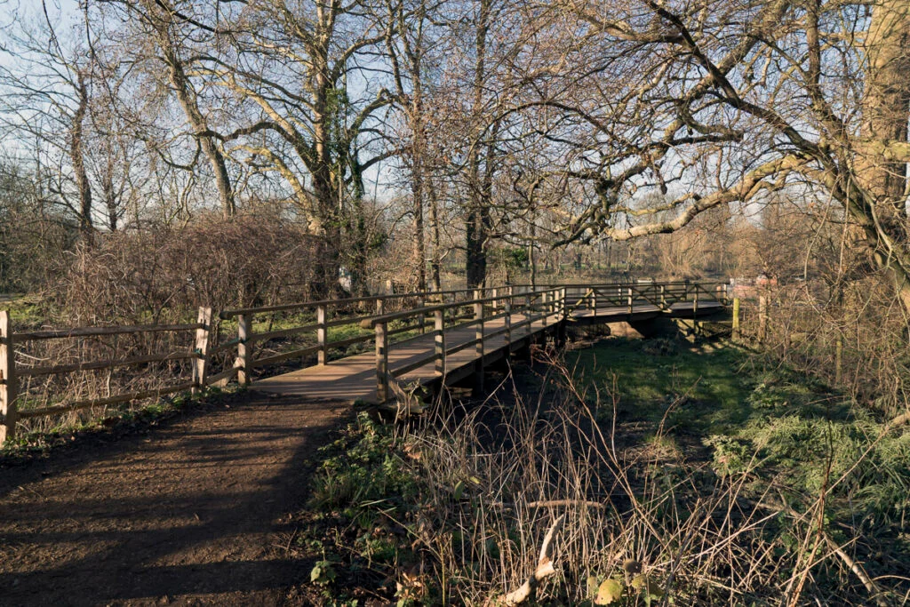 Eine hölzerne Fußgängerbrücke überquert an einem sonnigen Tag einen kleinen Bach in einem Park, der von blattlosen Bäumen und Unterholz umgeben ist.