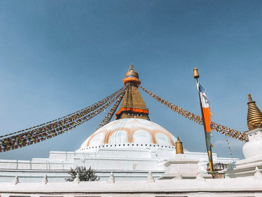 Ein großer weißer Stupa mit goldenem Turm und bunten Gebetsfahnen vor einem klaren blauen Himmel.