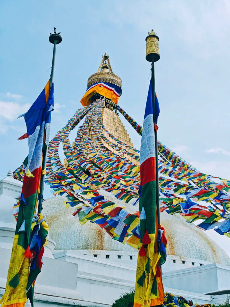 Ein großer Stupa mit Reihen bunter Gebetsfahnen, die von seinem Turm ausgehen, flankiert von zwei Fahnenmasten, unter klarem Himmel.