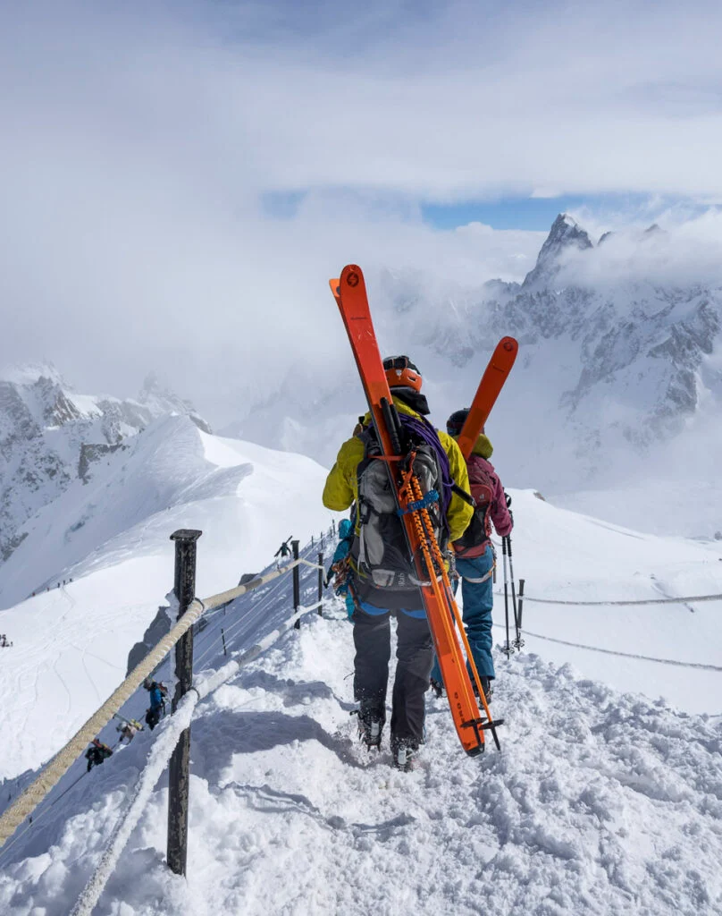 Zwei Skifahrer mit orangefarbenen Skiern wandern über einen schneebedeckten Bergrücken, umgeben von schneebedeckten Gipfeln unter bewölktem Himmel.