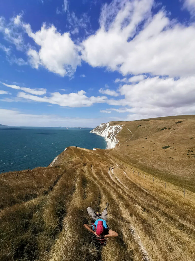 Eine Person liegt auf trockenem Gras mit Blick auf weiße Klippen und das Meer unter einem teilweise bewölkten Himmel.