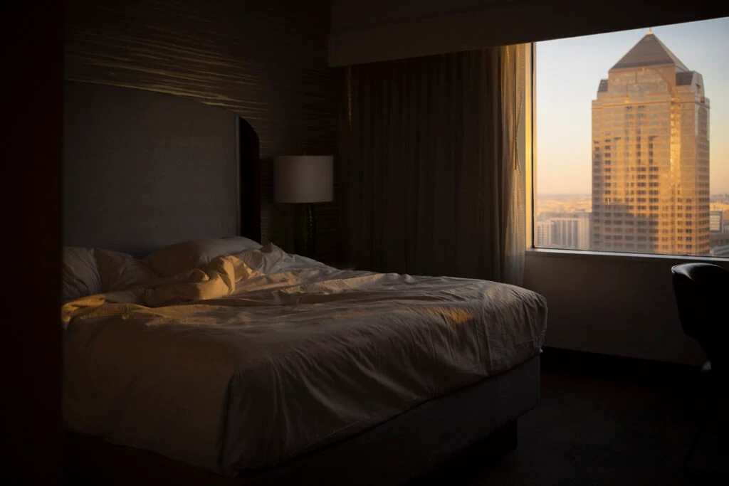 Ein Hotelzimmer mit einem gemachten Bett, beleuchtet von warmem Sonnenlicht, das durch ein Fenster mit Blick auf einen Wolkenkratzer in der Stadt strömt.