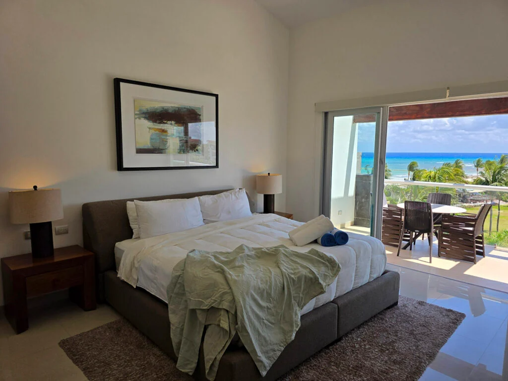 Ein Hotelzimmer mit einem Queensize-Bett, zwei Nachttischen und einer Glastür, die auf einen Balkon mit Meerblick führt.