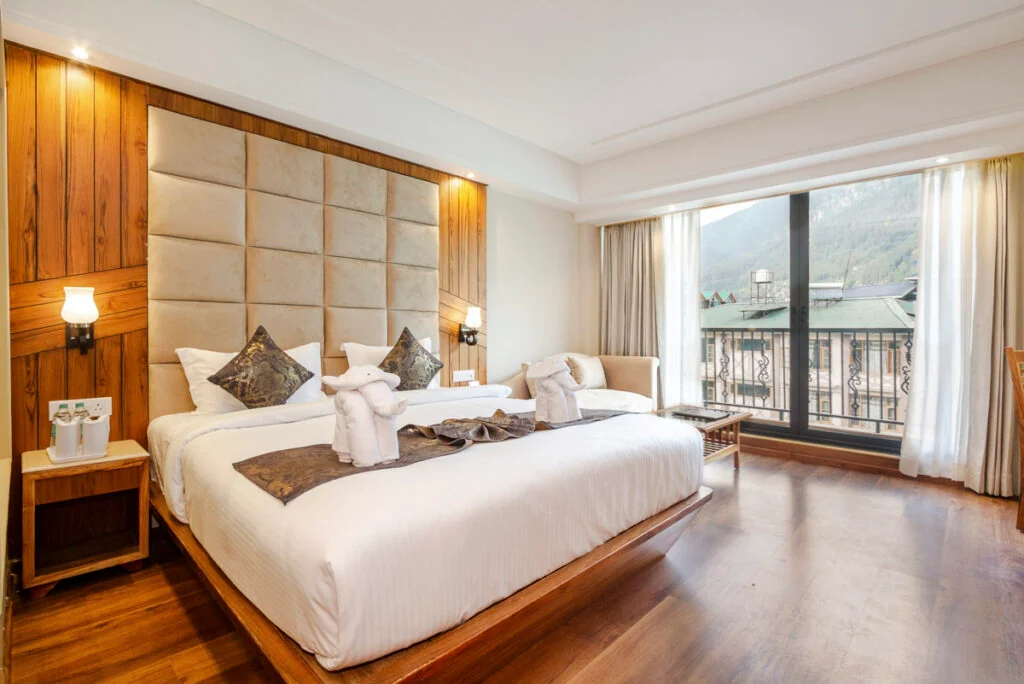 Hotelzimmer mit einem Kingsize-Bett mit zwei Handtuchelefanten, einem gepolsterten Kopfteil und einem großen Fenster mit Blick auf die Berglandschaft.