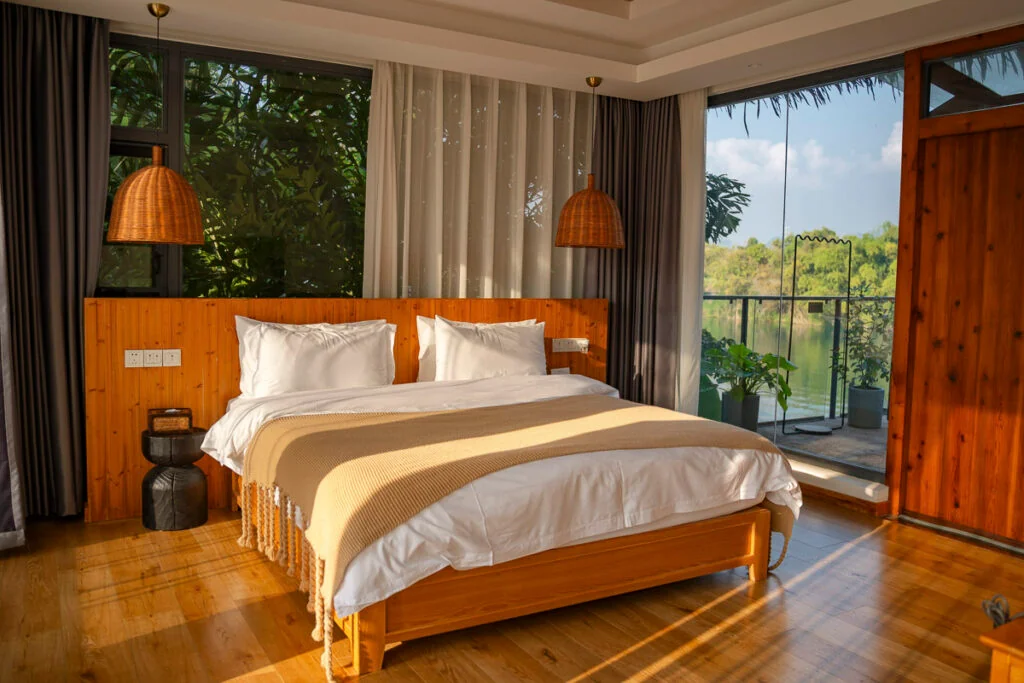 Ein Schlafzimmer mit einem Bettgestell aus Holz, weißer Bettwäsche und einer beigen Decke, vor einer Akzentwand aus Holz mit großen Fenstern und Blick auf einen See.
